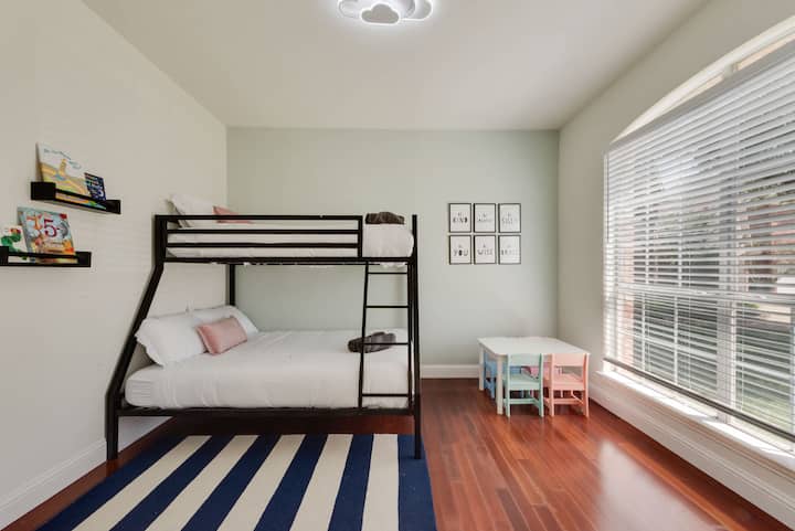 ห้องเด็กที่สนุกสนานและใช้งานได้ดี มีเตียงสองชั้นแบบเตียงเดี่ยวเหนือเตียงเต็ม ชั้นวางหนังสือ และโต๊ะสำหรับเด็กเล่นเกมหรือทำงานฝีมือ