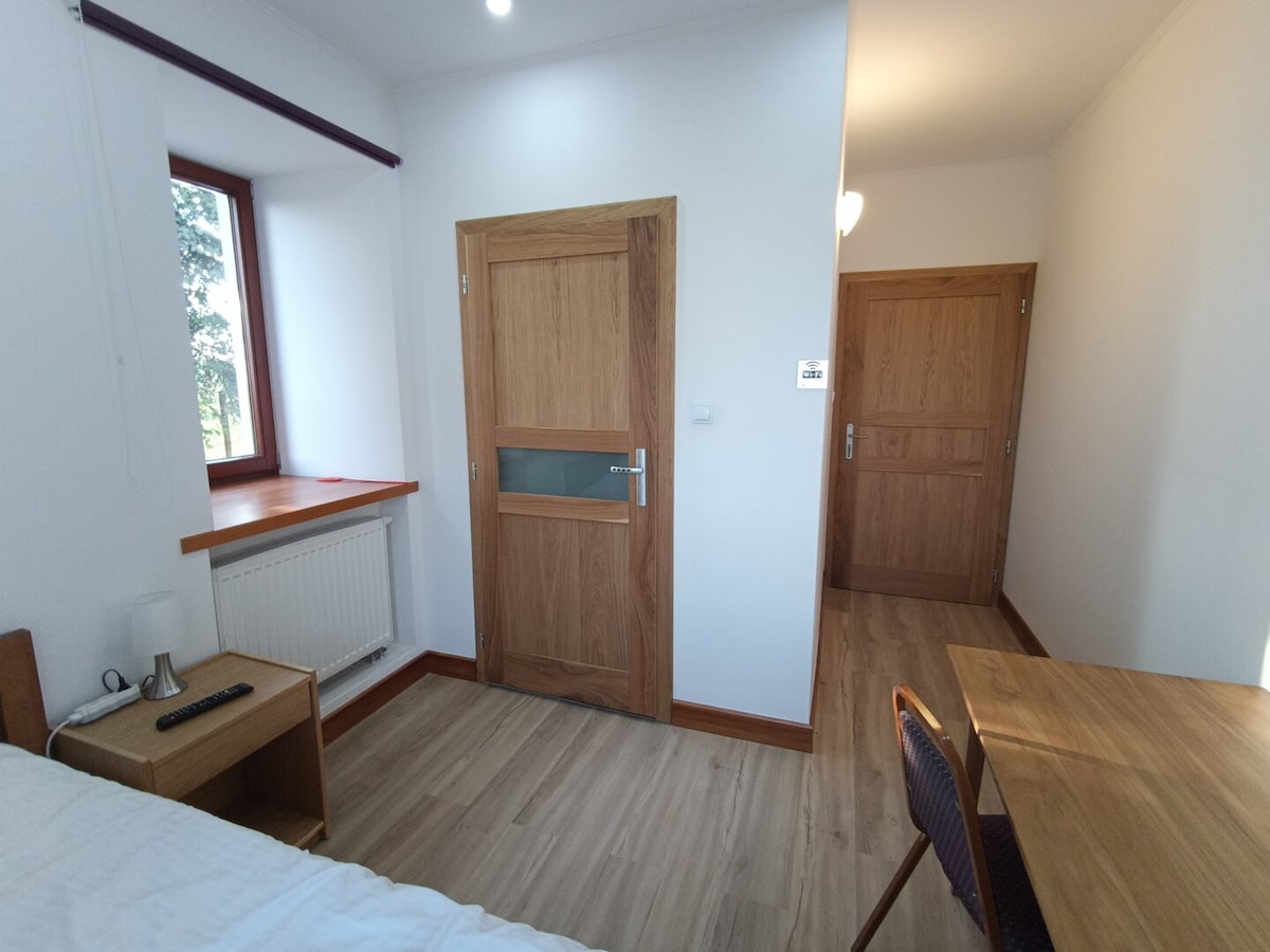 A bright single room is shown, featuring a large window that allows natural light to fill the space. A wooden desk and chair are situated near the window, and a door leads to an adjoining area. Light wooden flooring creates a warm atmosphere.