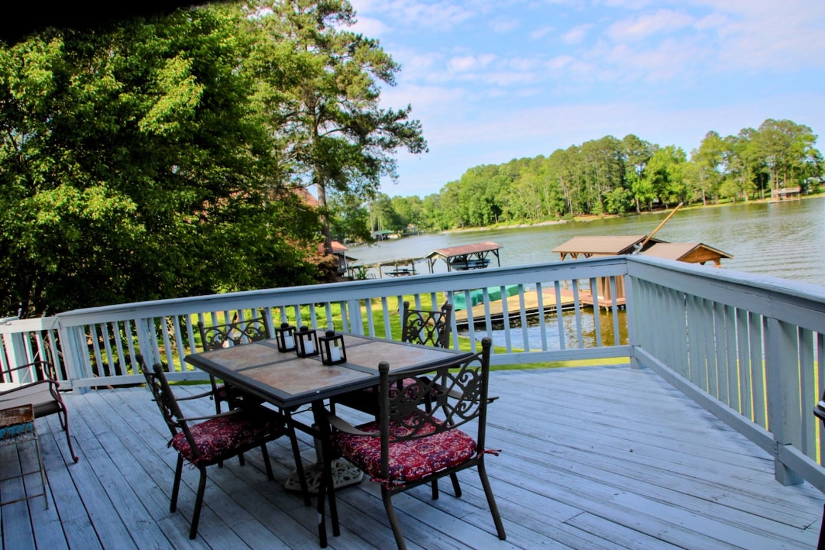 An outdoor deck offers a view of the lake, featuring a wooden table surrounded by four metal chairs with cushions. Lanterns are positioned on the table, with vibrant greenery and boathouses visible along the shoreline, creating a relaxed setting for gatherings.