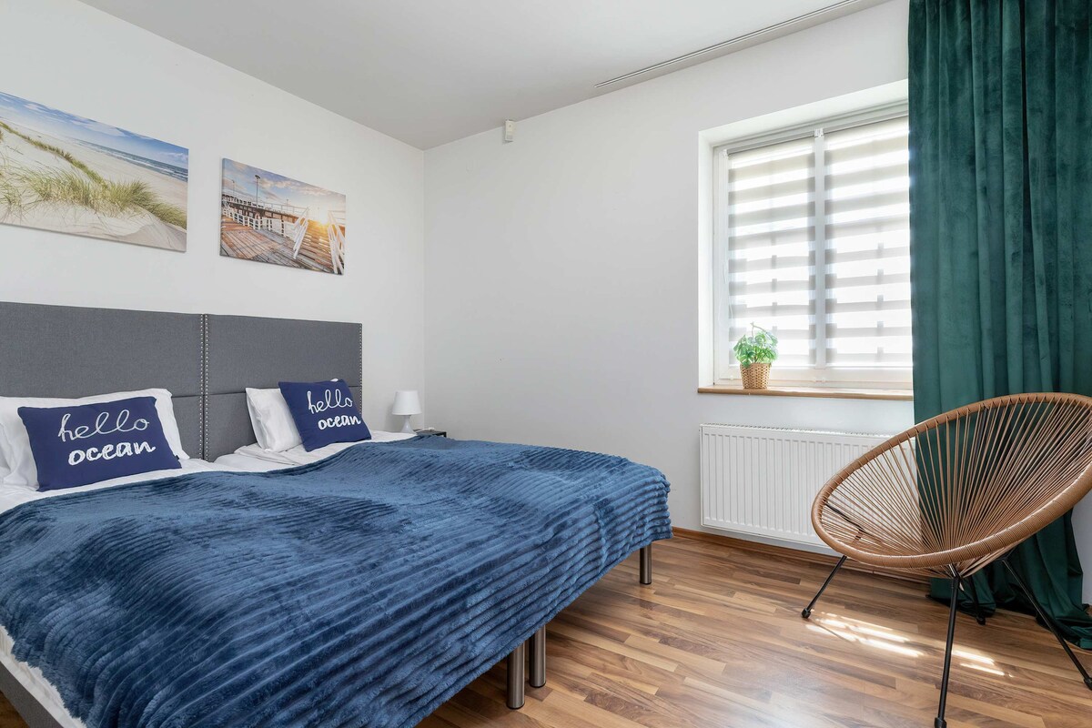 A spacious bedroom features a large bed with soft blue bedding and decorative pillows. Natural light filters through a partially covered window. A cozy armchair is positioned nearby, and beach-themed artwork adorns the walls, creating a calming atmosphere.