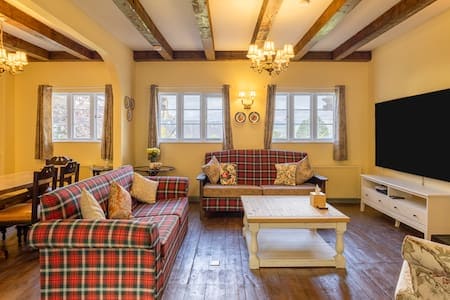 A welcoming living area features plaid sofas and a coffee table at its center. Natural light filters in through multiple windows, illuminating the rustic wooden beams above. A dining table with chairs is arranged in the background, creating a cozy space for gatherings.