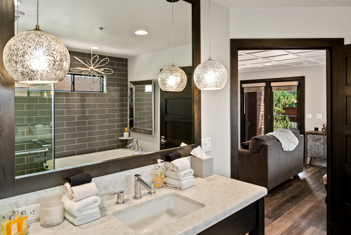 A custom bathroom is presented with a spacious vanity featuring quartz countertops and light fixtures adding a modern touch. Fluffy bath towels are neatly arranged beside bath products, while a freestanding tub is visible in the background. The design offers a seamless view into the adjoining living area.