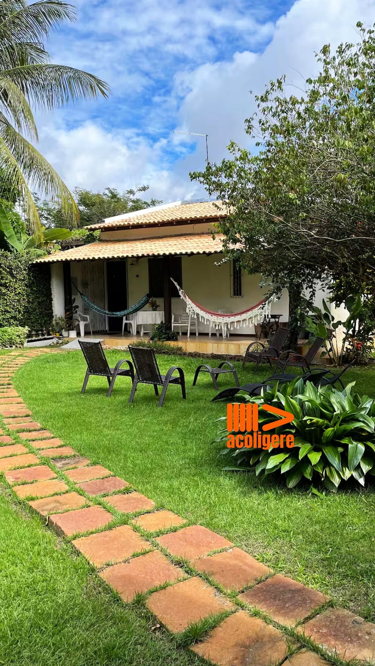 A well-maintained garden is featured in front of a cozy cottage. Lush green grass surrounds a stone pathway leading to a covered porch. Several patio chairs are arranged under the shade of trees, complemented by hammocks. The clear sky adds a bright ambiance to the scene.