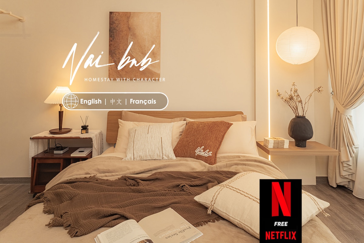 The bedroom features a spacious bed adorned with neutral-toned linens and decorative pillows. A simple bedside table holds a lamp, while soft lighting enhances the calming atmosphere. Large windows draped with sheer curtains allow for natural light, creating a serene retreat.