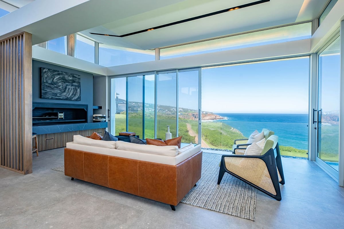 A modern living area features a light-colored sectional sofa and an accent chair, positioned to offer expansive ocean views through large glass walls. The room is framed by natural light, highlighting the sleek design and inviting a seamless indoor-outdoor experience.