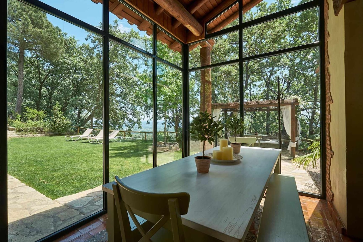 An airy dining area is enclosed by large glass walls, allowing views of the surrounding greenery. A wooden table is set with decorative candles, while outdoor lounge chairs are visible on the manicured lawn beyond. Natural light floods the space.