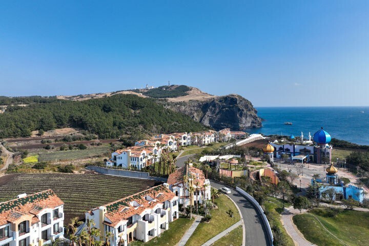 An aerial view of a coastal resort area showcases white buildings with terracotta roofs surrounded by lush greenery. A winding road runs through the property, leading towards a vibrant dome structure near the shoreline with waves gently lapping against the rocky coast.