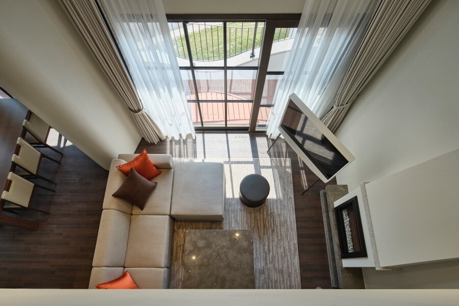 A spacious living area features large windows draped with sheer curtains, allowing natural light to fill the space. A light-colored sectional sofa is complemented by orange accent pillows. A coffee table sits on a textured rug, with a modern flat-screen television mounted on the wall.