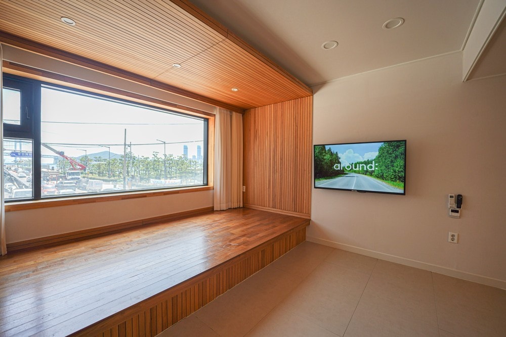 A spacious living area features a large window with a view of the outdoor scenery, complemented by wooden accents on the walls and flooring. A flat-screen television is mounted on one wall, providing entertainment options. Natural light fills the room, creating an open atmosphere.