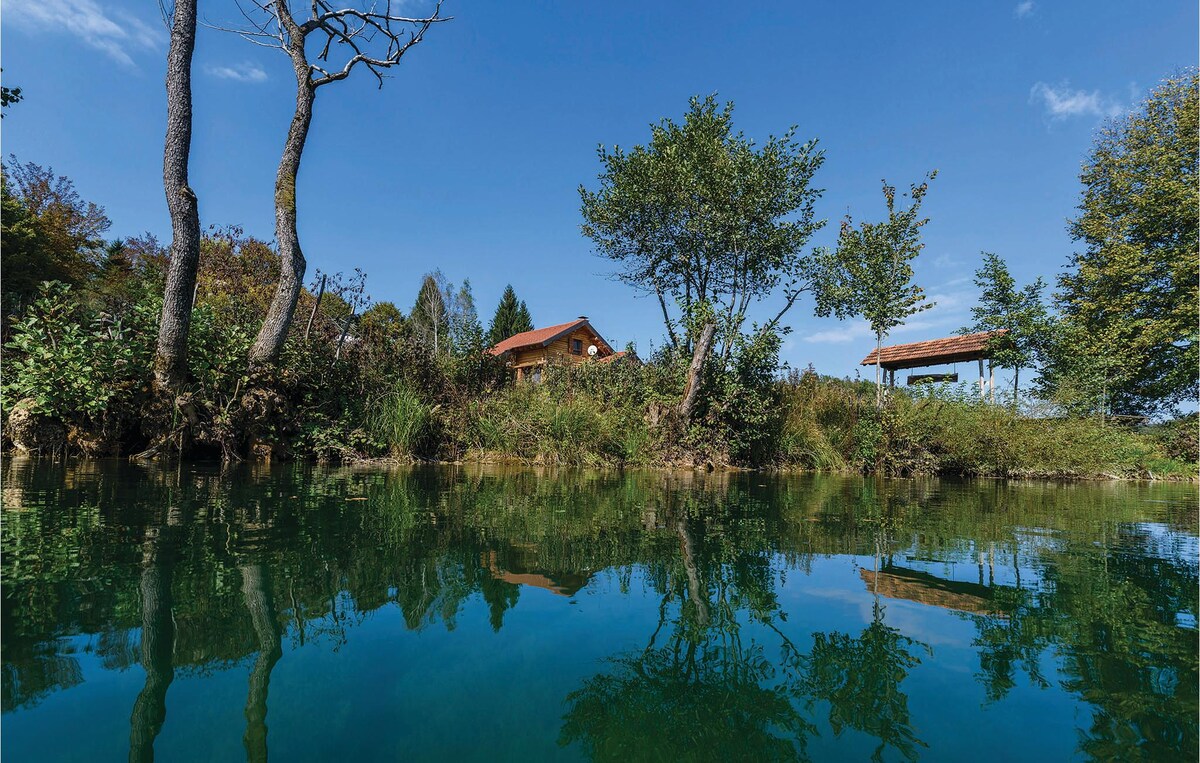 The tranquil scene displays a rustic house surrounded by greenery, reflected in the calm water. The setting includes trees and a small gazebo nearby, all under a clear blue sky. The natural landscape suggests a serene environment for relaxation.