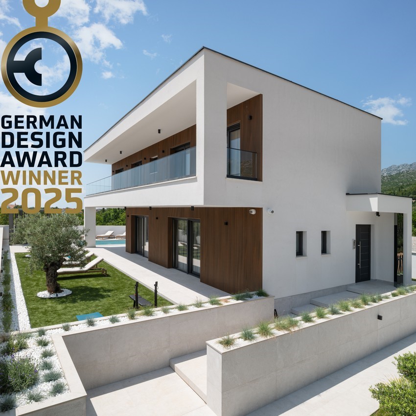 The modern exterior of Villa Cvita showcases clean lines and a harmonious blend of white and natural wood elements. Lush greenery surrounds the property, enhancing its contemporary design. A German Design Award badge highlights the villa's architectural excellence, set against a clear blue sky.