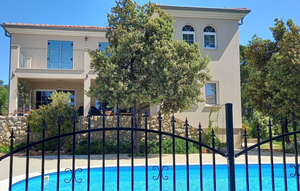 A modern villa is seen from the garden, with lush greenery framing the exterior. The private swimming pool, showcased in vibrant blue, complements the stone details of the property. Large windows and a balcony enhance the overall inviting appearance of the home.
