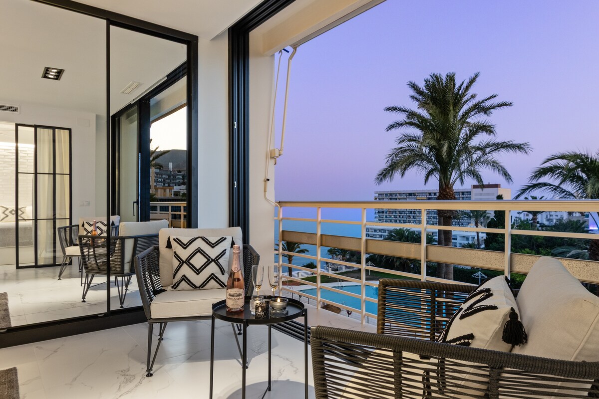An outdoor terrace area is displayed, featuring modern seating with decorative cushions. A small table holds two glasses, accompanied by a bottle. Large sliding windows open to reveal a view of palm trees and the evening sky, enhancing the tranquil ambiance.