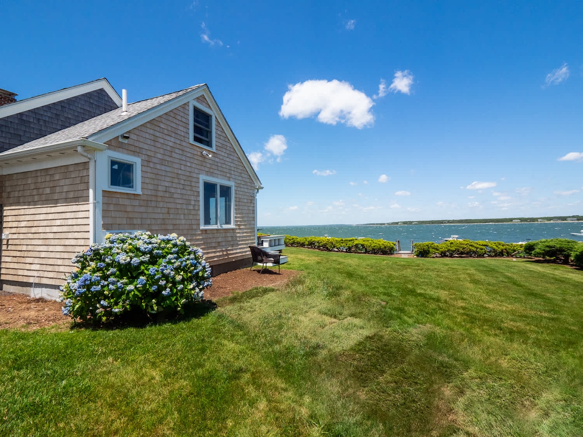 The exterior of the oceanfront home showcases a well-maintained yard with lush green grass and blooming hydrangeas. The shingles of the house reflect coastal charm, while panoramic views of Buzzards Bay extend in the background, under a clear blue sky adorned with a few fluffy clouds.