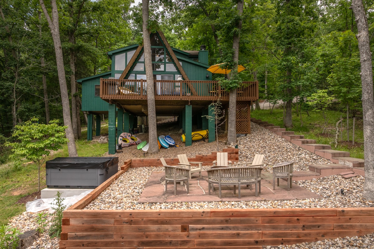The property features a two-story chalet nestled among trees, with a large deck overlooking a landscaped outdoor area. A seating arrangement with multiple chairs surrounds a fire pit on a stone patio, and colorful water toys are stored nearby.