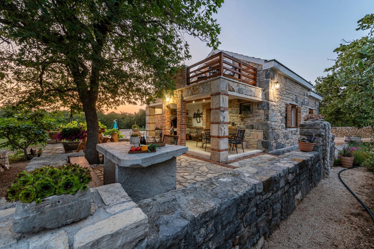 The rustic villa exterior features stone walls, a spacious terrace with wooden railings, and decorated with various potted plants. A stone barbecue area is visible, surrounded by a garden with olive trees. Soft lighting enhances the warm ambiance as evening sets in.