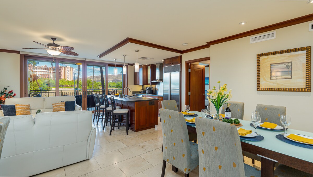 An open-concept living area presents a spacious dining table set with dishes and glasses. A modern kitchen, equipped with stainless steel appliances, features a large island with additional seating. Large windows allow natural light to fill the space, offering views of the outdoors.