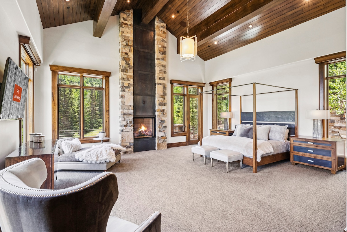 the massive primary suite with a fireplace that stretches to the ceiling, and a 4 poster bed with cozy seating and views from 3 walls with tall windows and mountain views