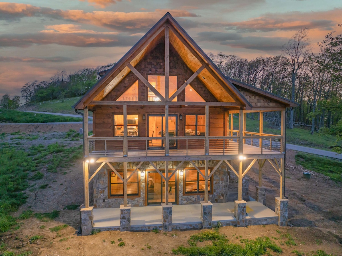 A newly constructed mountain retreat is showcased with a large deck and expansive windows. The structure's wooden beams and stone accents highlight its rustic charm, while surrounding greenery and a soft sunset sky add to the inviting scenery.