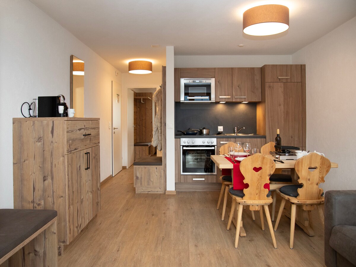 The open-plan living area showcases a modern kitchen equipped with stainless steel appliances, including an oven and stovetop. A wooden dining table with chairs is positioned nearby. Natural light fills the space, highlighted by warm pendant lighting and light-colored wood flooring.