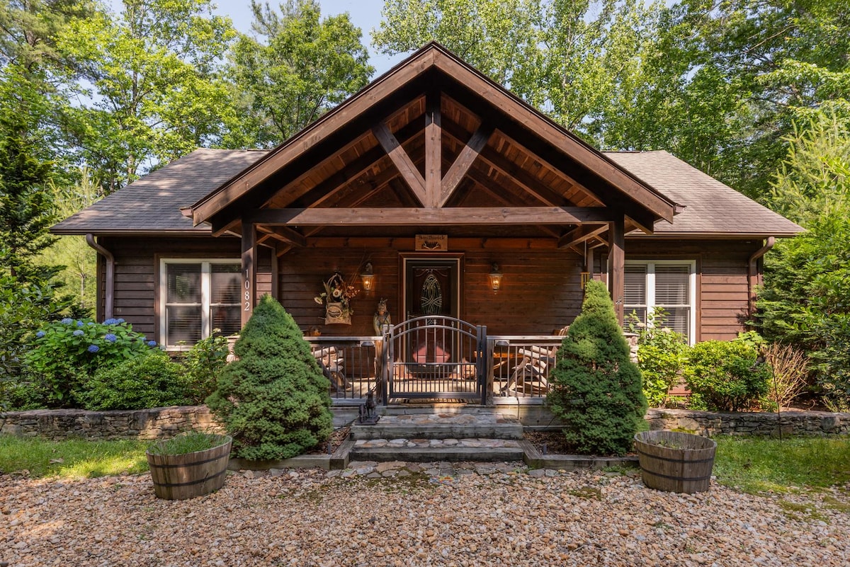 A welcoming cabin entrance is framed by two lush green shrubs and decorative wooden planters. The front porch features a wooden railing, leading to the main door, which is adorned with rustic accents under a triangular roof supported by wooden beams.