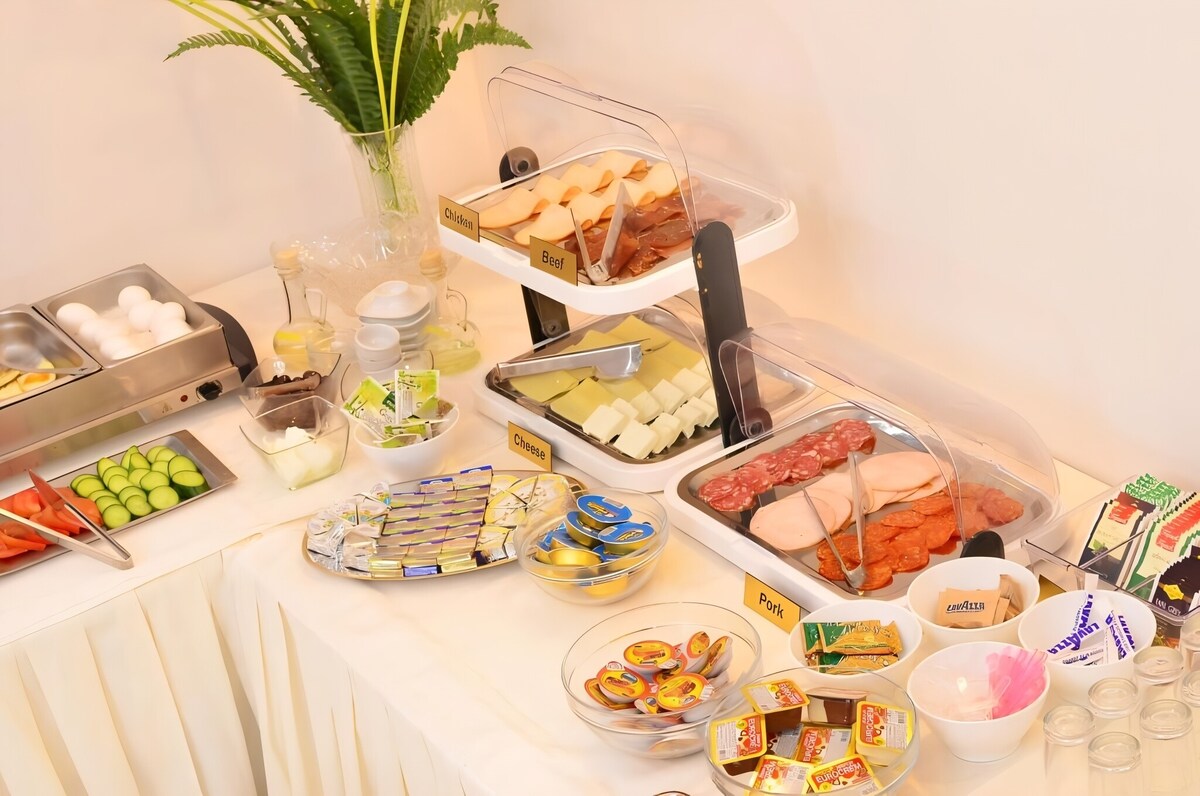 A breakfast buffet is displayed with a variety of food items. Cold cuts, cheeses, and vegetables are arranged on platters. A selection of drinks and spreads is presented in labeled containers, along with individual servings of snacks and desserts for guests to enjoy.