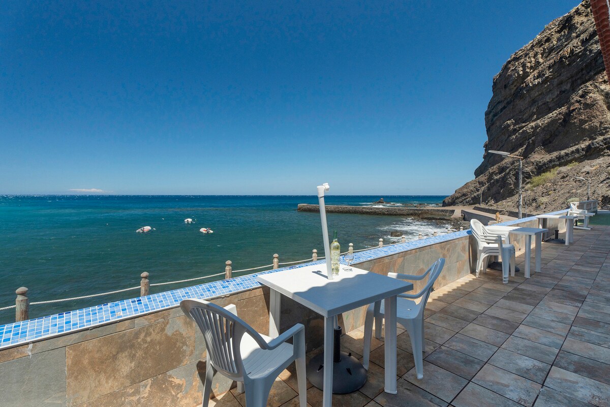 A coastal view showcases the ocean meeting the rocky shore, with clear blue waters stretching toward the horizon. Several white chairs are positioned around tables along a tiled patio, providing an inviting space for relaxation against the backdrop of the mountainous terrain.