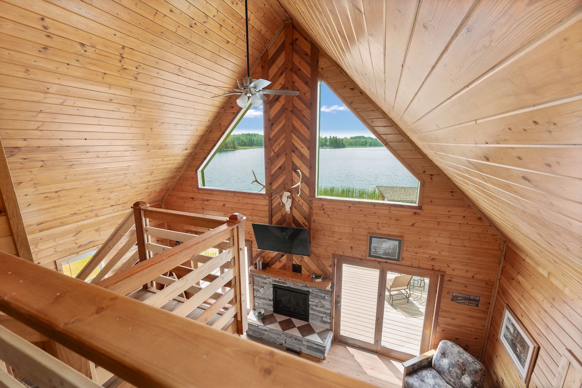 The interior of the cabin features high wood-paneled ceilings and large triangular windows that offer a view of the serene lakeside. A stone fireplace is positioned beneath a mounted television, and a comfortable seating area invites relaxation.