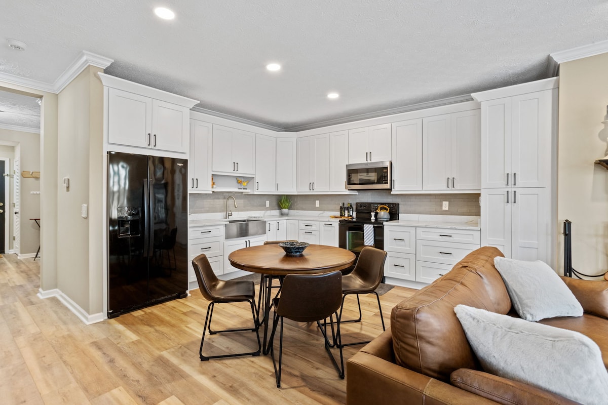 A modern kitchen and dining area is displayed, featuring sleek white cabinetry and a dark refrigerator. A round wooden table with four chairs occupies the center, while a cozy seating area with a leather sofa is visible in the foreground, creating a welcoming space for relaxation and dining.