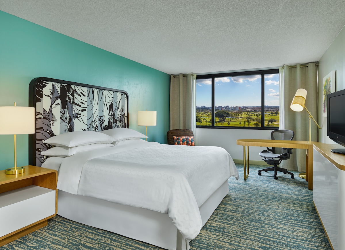 A well-appointed hotel room features a spacious king bed with decorative bedding. A large window offers views of the outside landscape. A comfortable chair and desk are included, alongside a flat-screen television. The walls are painted in a calming teal shade, adding to the room's bright ambiance.