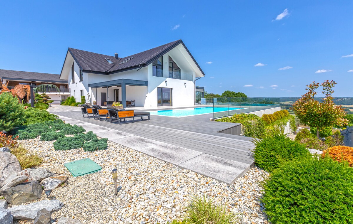 An elegant property is showcased with a spacious outdoor terrace and a shimmering pool. Multiple lounge chairs are arranged beside the water, creating an inviting space for relaxation. Lush greenery and manicured gardens enhance the landscape, with gentle hills visible in the background.