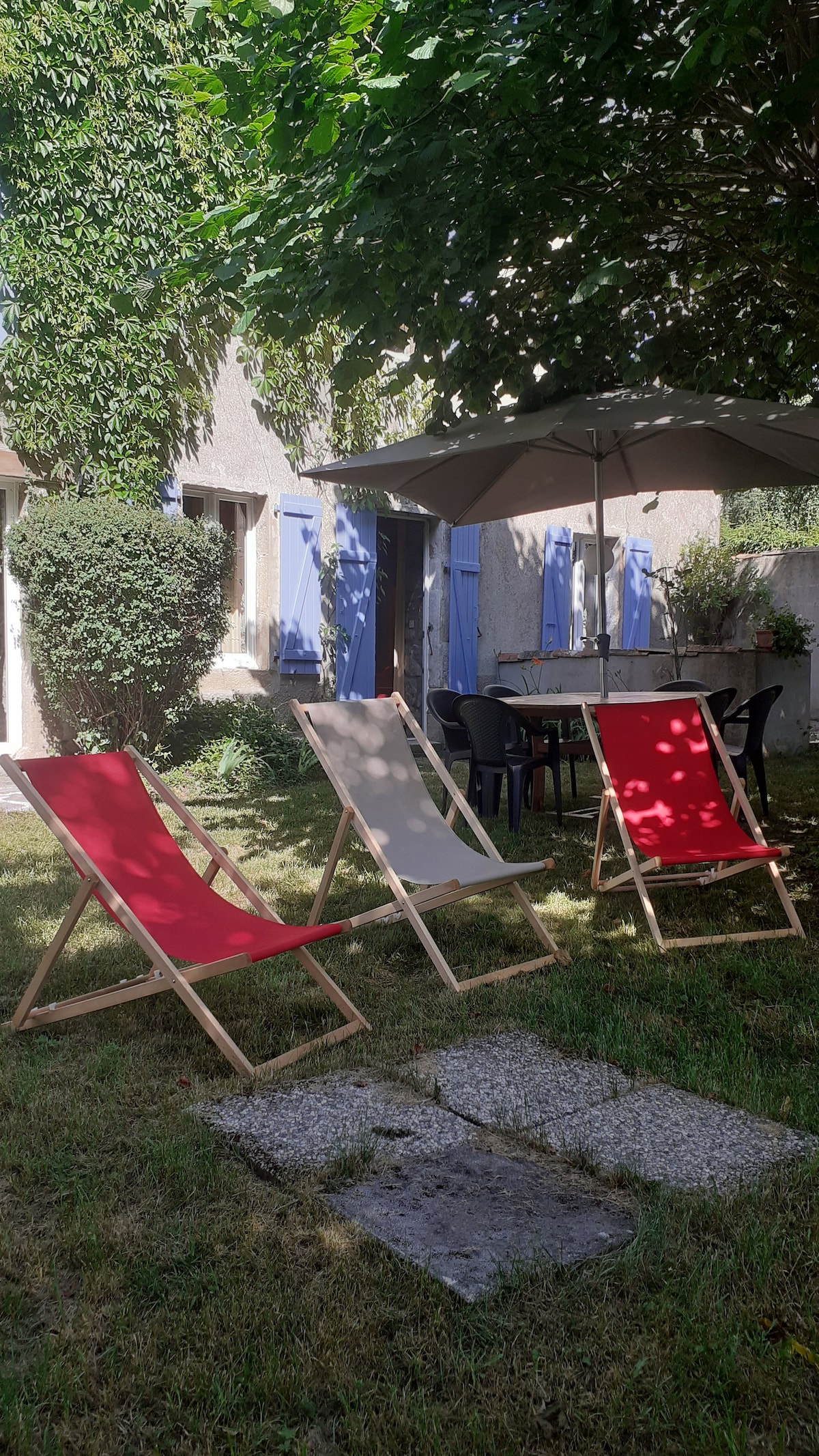A private outdoor space features three lounge chairs in red and beige, arranged on the grass under the shade of a tree. A patio umbrella provides cover for a nearby dining table surrounded by black chairs. The residence is visible in the background with blue shutters.
