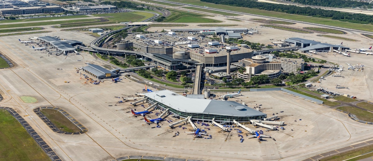 An aerial view of Tampa International Airport showcases multiple parking areas and terminal buildings. The expansive runway is visible, lined with parked planes and various ground service vehicles. Surrounding green landscapes provide a contrasting backdrop, highlighting the airport's extensive layout.