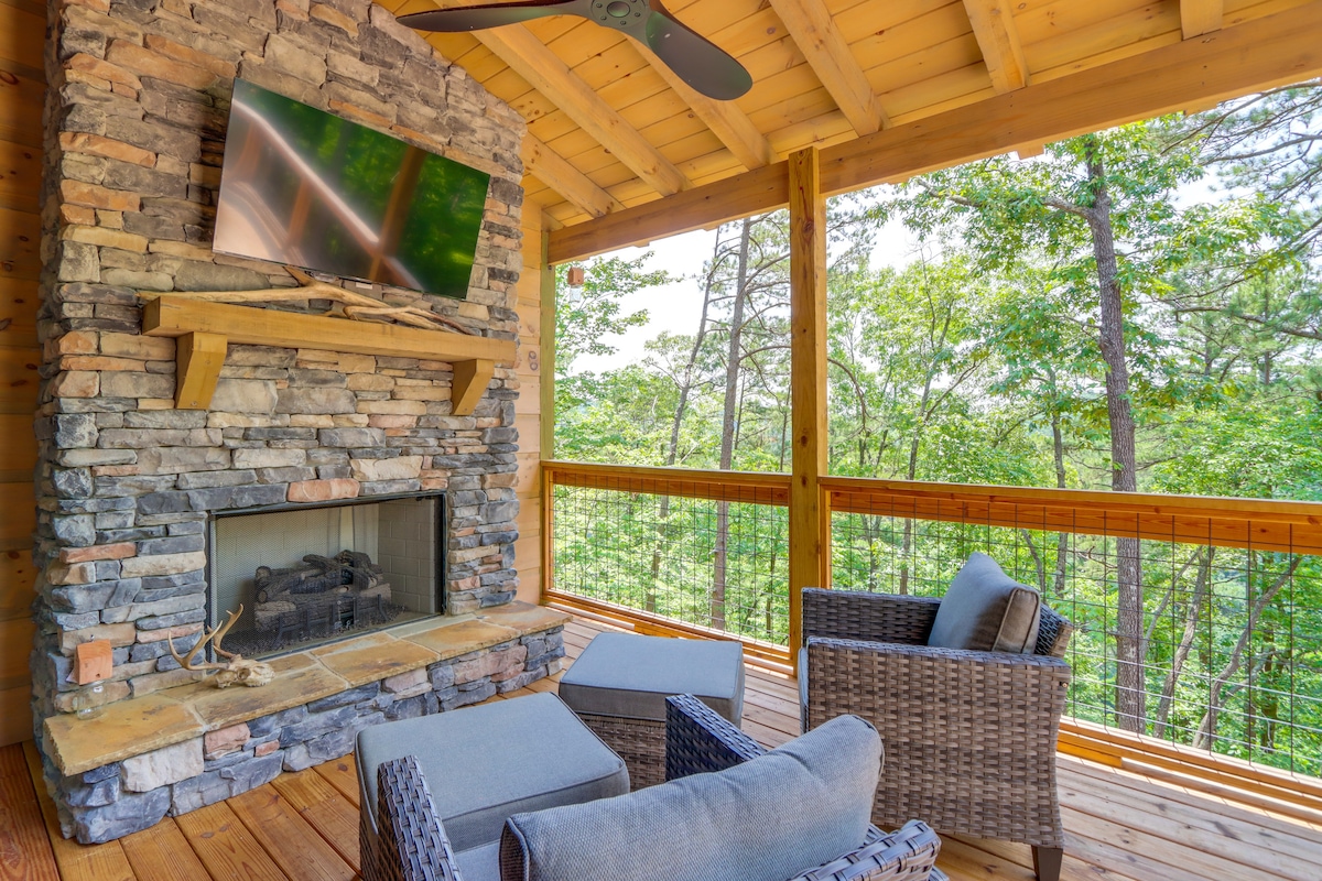 A covered deck area features a stone fireplace with a mounted television overhead. Comfortable seating includes two woven chairs with cushions, positioned to enjoy the surrounding wooded views. Sunlight filters through the trees, creating a serene outdoor atmosphere.