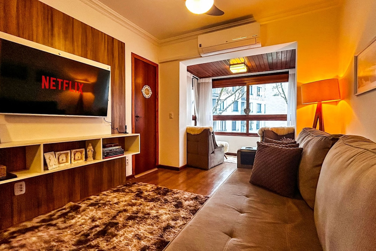 A comfortable living area features a plush sofa positioned opposite a wall-mounted Smart TV, displaying the Netflix logo. Natural light enters through large windows, creating a warm ambiance. A cozy reading nook is visible, along with a stylish floor lamp and decorative accents.