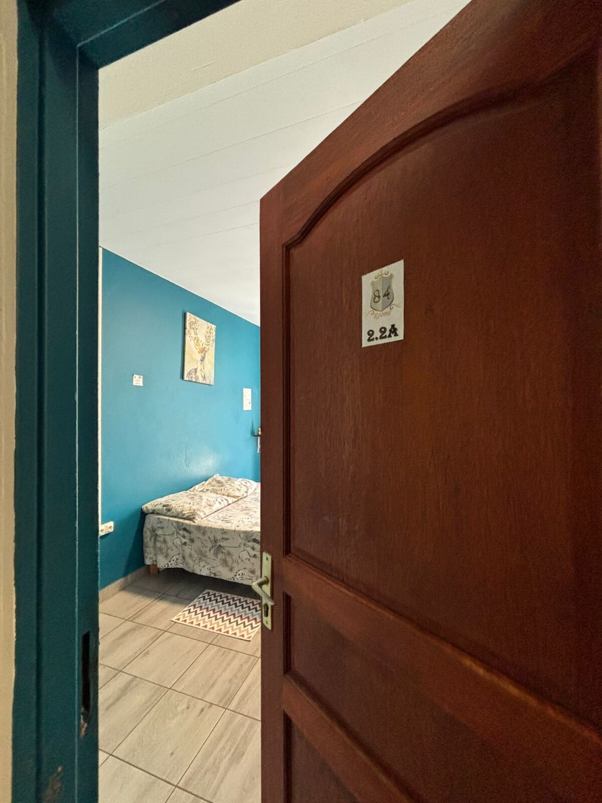 A view into a private room is presented through an open door. The room features a bed covered with a patterned blanket against a teal wall. Light falls onto the tiled floor, illuminating details of a small rug positioned near the foot of the bed.