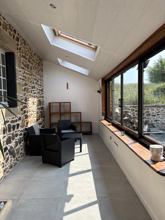 A bright, sunlit veranda is showcased, featuring natural light from skylights and large windows. Two black chairs and a wooden shelf add functional decor, while the stone wall provides a rustic contrast. A small white plant pot sits on the ledge, enhancing the serene environment.