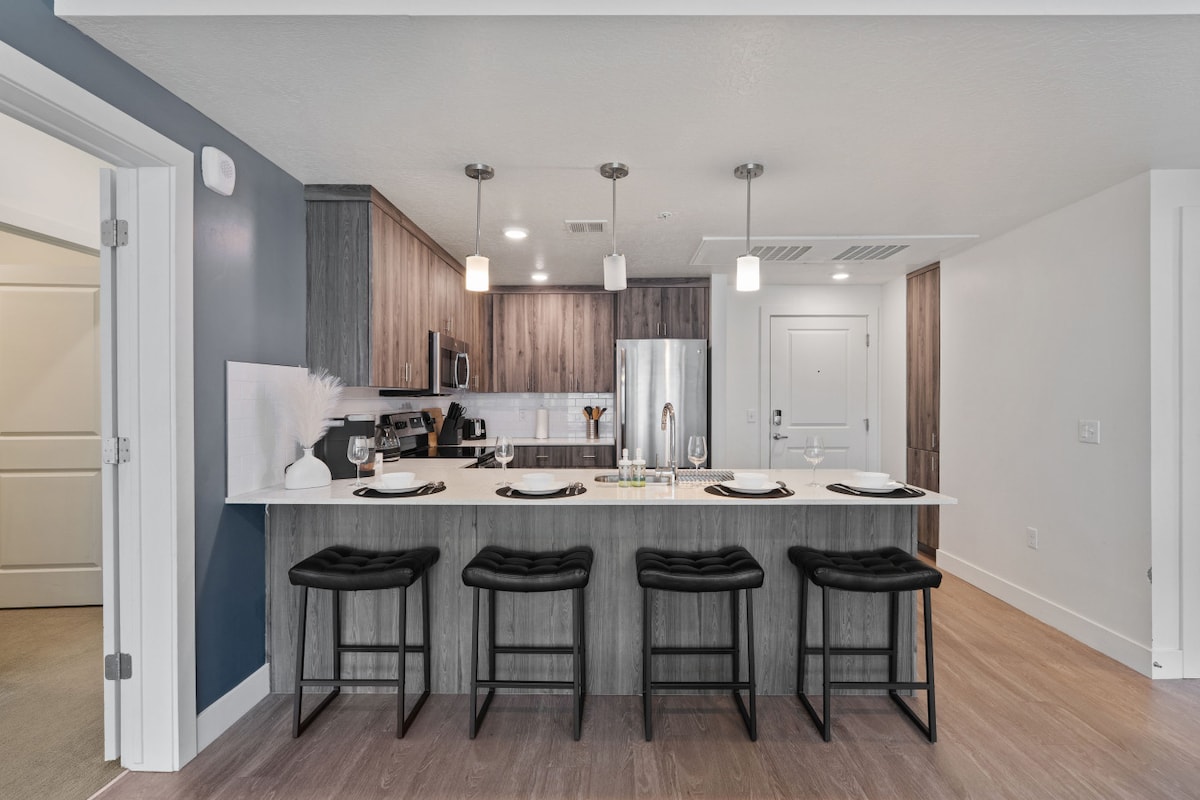 A modern kitchen island is highlighted, featuring a spacious countertop and sleek stainless steel appliances. Four cushioned barstools are arranged neatly, and various kitchen utensils are placed on the counter. Soft lighting accents the warm wood cabinetry and the neutral color scheme.