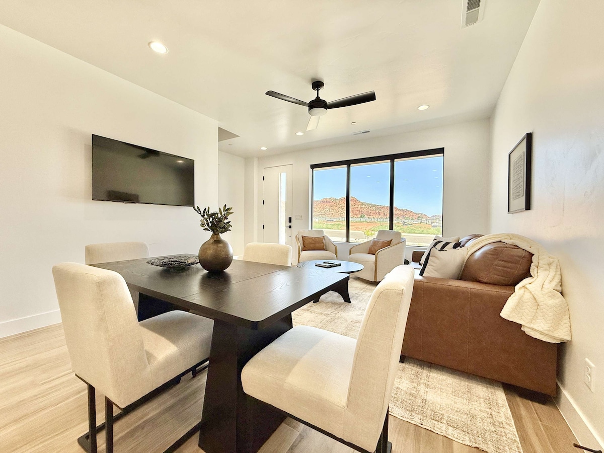 A modern living and dining space features a dark wooden table surrounded by four upholstered chairs. A brown leather couch sits against the wall, complemented by colorful throw pillows. Large windows showcase views of the surrounding red rock landscape, while a wall-mounted TV is visible.