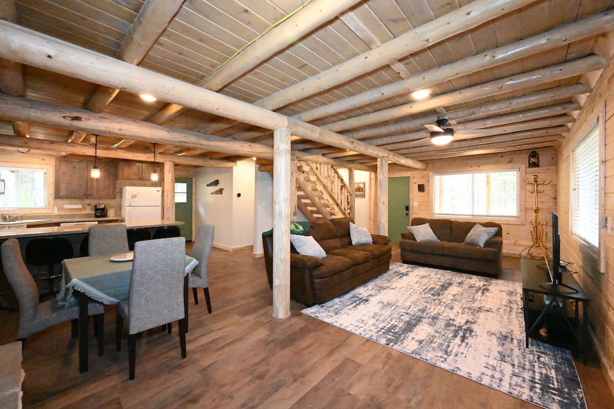 A spacious living area features rustic log beams and a cozy seating arrangement with two couches. A dining table is positioned nearby, accompanied by chairs. Light enters through window treatments, illuminating a patterned area rug. Stairs lead to an upper level, enhancing the room's open feel.