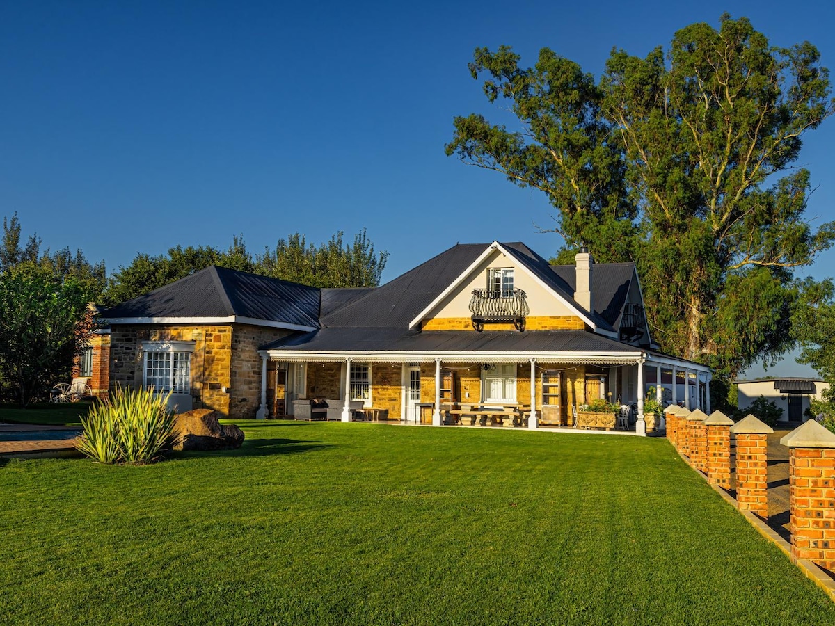The exterior of a spacious family home is showcased, with a well-manicured lawn in the foreground. The structure features a combination of stone and wood accents, while multiple seating areas are visible on the porch, creating inviting spaces for relaxation.