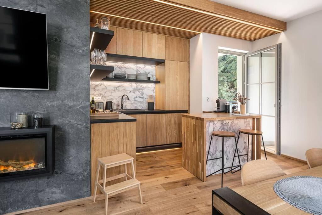 A modern kitchen space is showcased with natural wood cabinetry and marble accents. A cozy fireplace is integrated into the living area. A small breakfast bar sits adjacent to the kitchen, complemented by a wooden step stool. Light streams in through a glass door leading to an outdoor area.