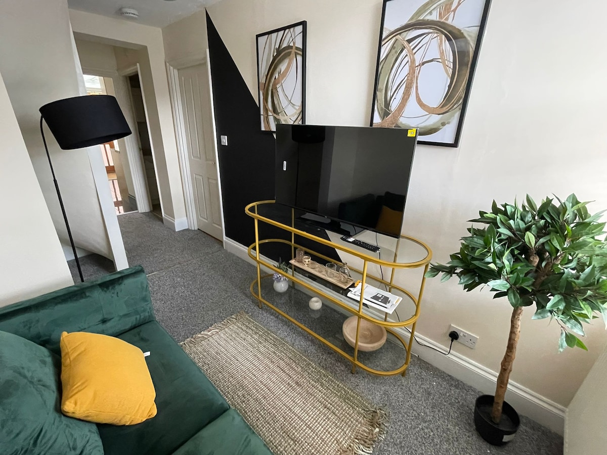A separate living room area features a green sofa adorned with a yellow cushion. A sleek, yellow console table displays decorative items beneath a smart TV. A potted plant adds a touch of greenery, while a textured area rug defines the sitting space.