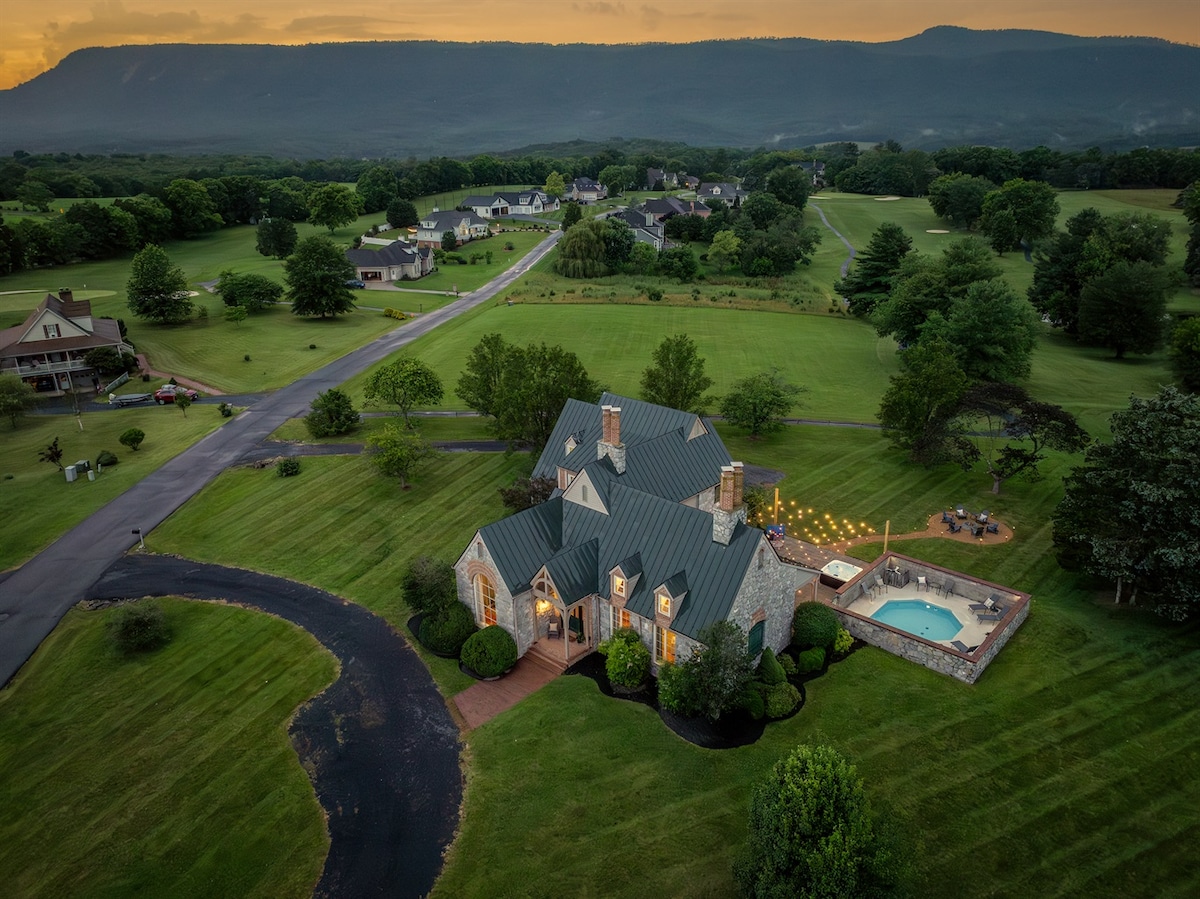 An aerial view captures the elegant stone home with a distinctive roof, set on a spacious lot surrounded by lush greenery. The private pool is visible in the backyard, complemented by string lights, with the scenic mountain range providing a serene backdrop.