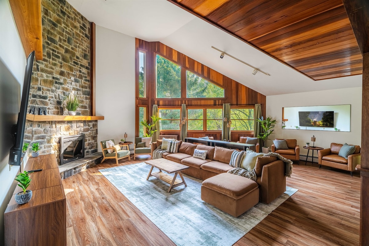 A spacious living area features soaring ceilings and a mix of wooden tones. Large windows provide ample natural light and views of the surrounding forest. Comfortable seating is arranged around a low coffee table, with a gas fireplace enhancing the cozy ambiance.