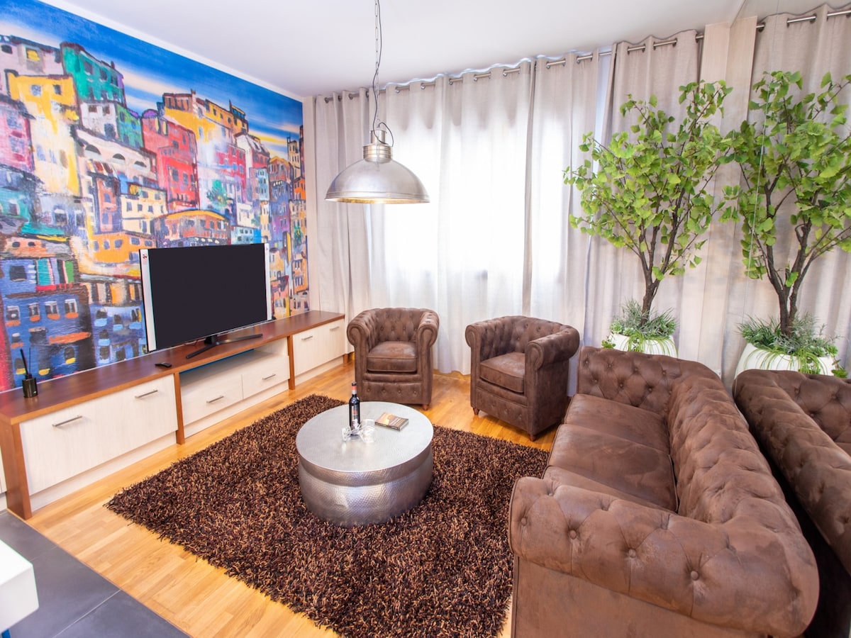 A bright living area features a large art mural of colorful houses, complemented by two comfortable armchairs and a spacious sofa. A round coffee table sits in front of the sofa, all placed over a textured and inviting rug. Soft natural light filters through the sheer curtains.