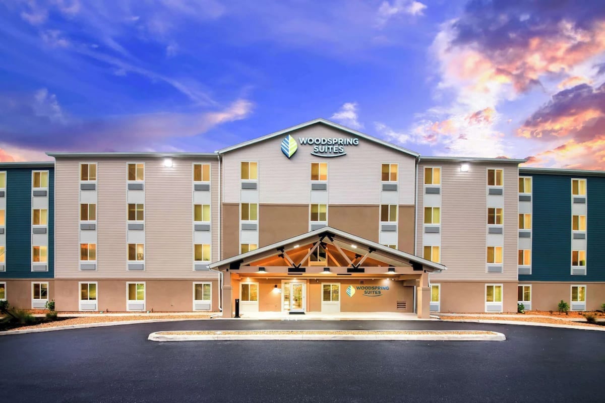 The exterior of WoodSpring Suites is shown, featuring a modern three-story building with a welcoming entrance. Large windows reflect the soft evening light. The architectural design includes a blue and beige color scheme, accented by wooden elements at the entrance.