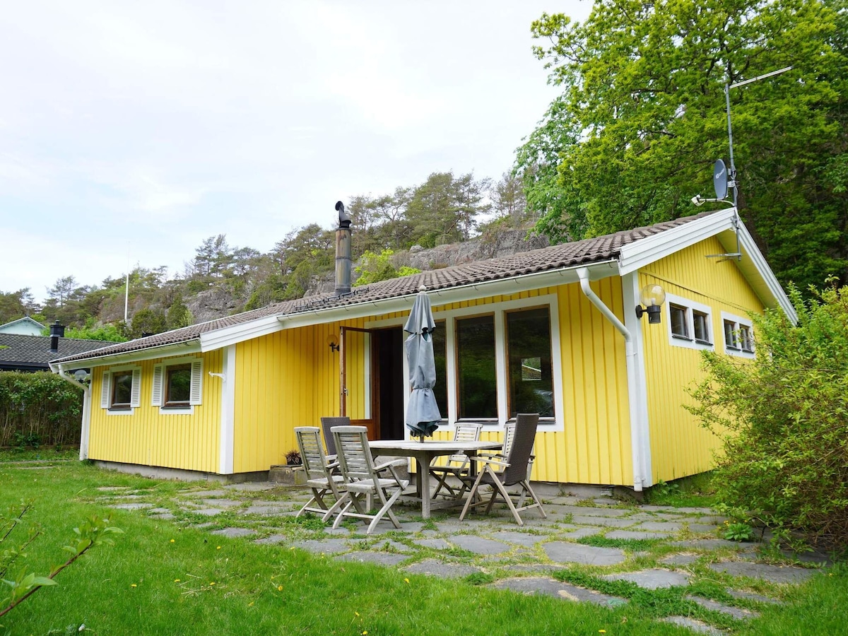 A bright yellow cottage is set against a backdrop of lush greenery and rocky hills. The spacious patio features an outdoor dining table with six chairs and an umbrella, surrounded by well-maintained grass. Large windows provide natural light and views of the surrounding nature.