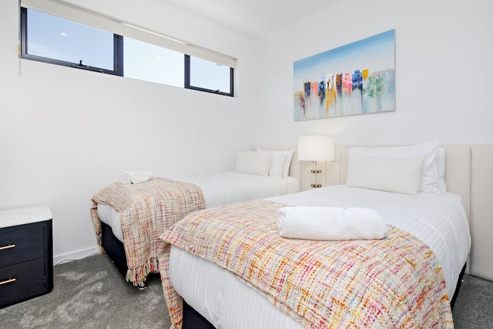 שתי מיטות יחיד עם שמיכות בהירות ויצירות אמנות של נופים חוף שמכניסות אתכם לאווירה.