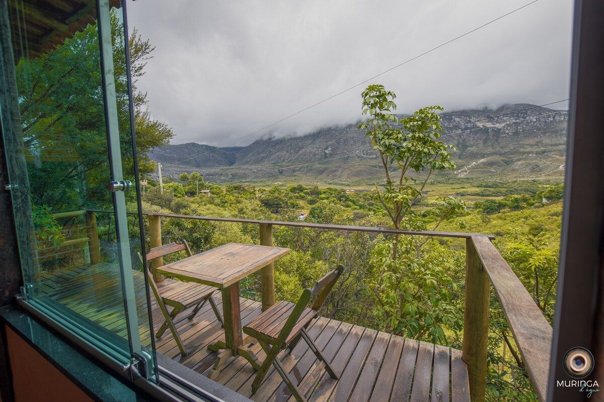A wooden deck is visible from the interior, featuring a table and chairs. Lush green foliage surrounds the area, offering a view of the distant mountains shrouded in mist. The tranquil landscape provides a peaceful atmosphere.
